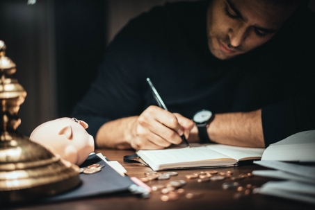 A man in a dark room writes in a notebook next to a piggy bank